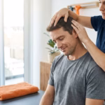 Therapist massages a man's scalp and neck from behind in a bright therapy room.